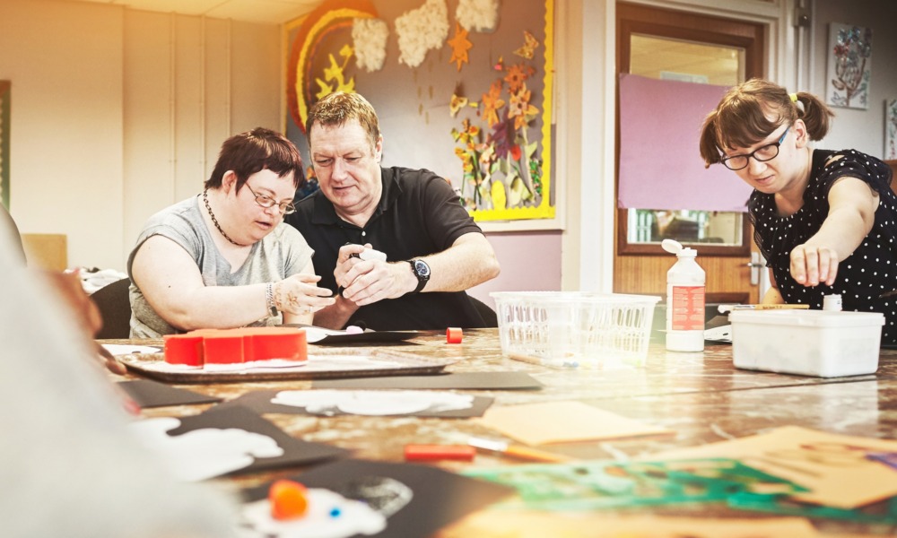disability-informed mentorship for youth with disabilities should be a right, not a matter of chance_feature: an older man helps a young disabled person at table with task while another disabled person works on their own