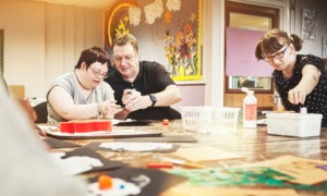 disability-informed mentorship for youth with disabilities should be a right, not a matter of chance_feature: an older man helps a young disabled person at table with task while another disabled person works on their own