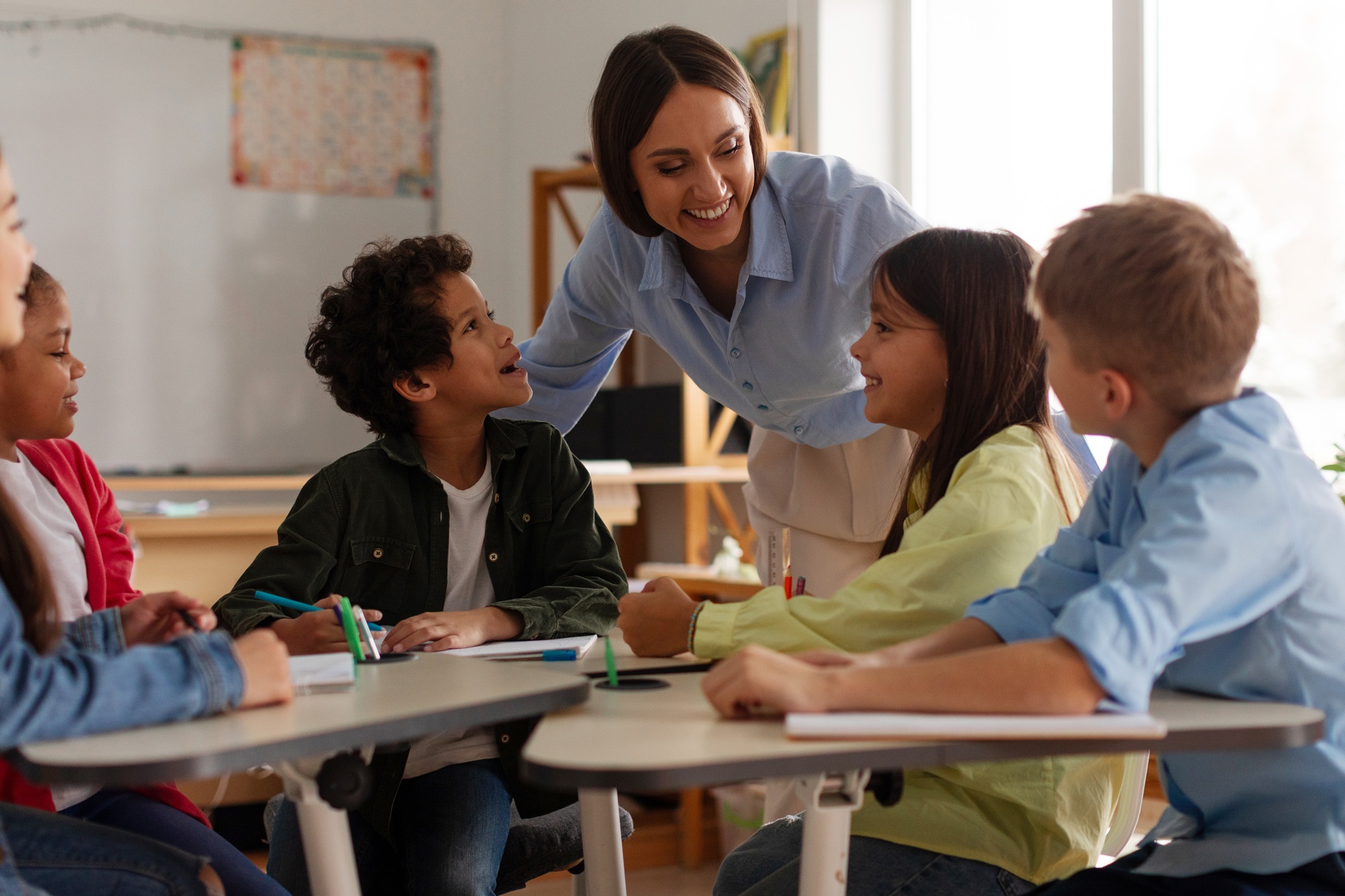 More than a test-How trauma can disrupt learning_feature: young female teacher helps kids in their desks while they smile up at her