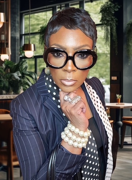 Sharon McDaniel headshot: black woman wearing striped blazer with short hair and thick, dark rimmed glasses with chin on hand with restaurant setting in background