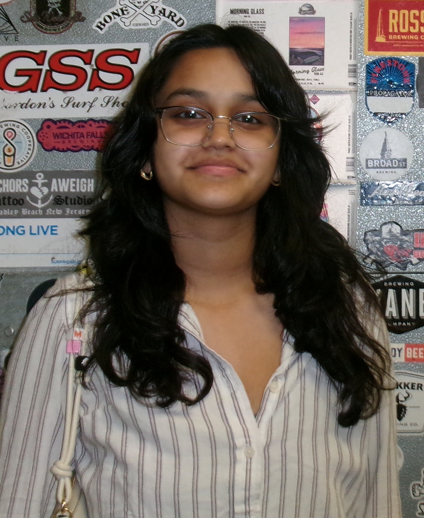 Tanisi Sahai headshot: teen girl of Indian descent standing in front of wall of stickers wearing glasses and vertical-striped collared shirt and glasses