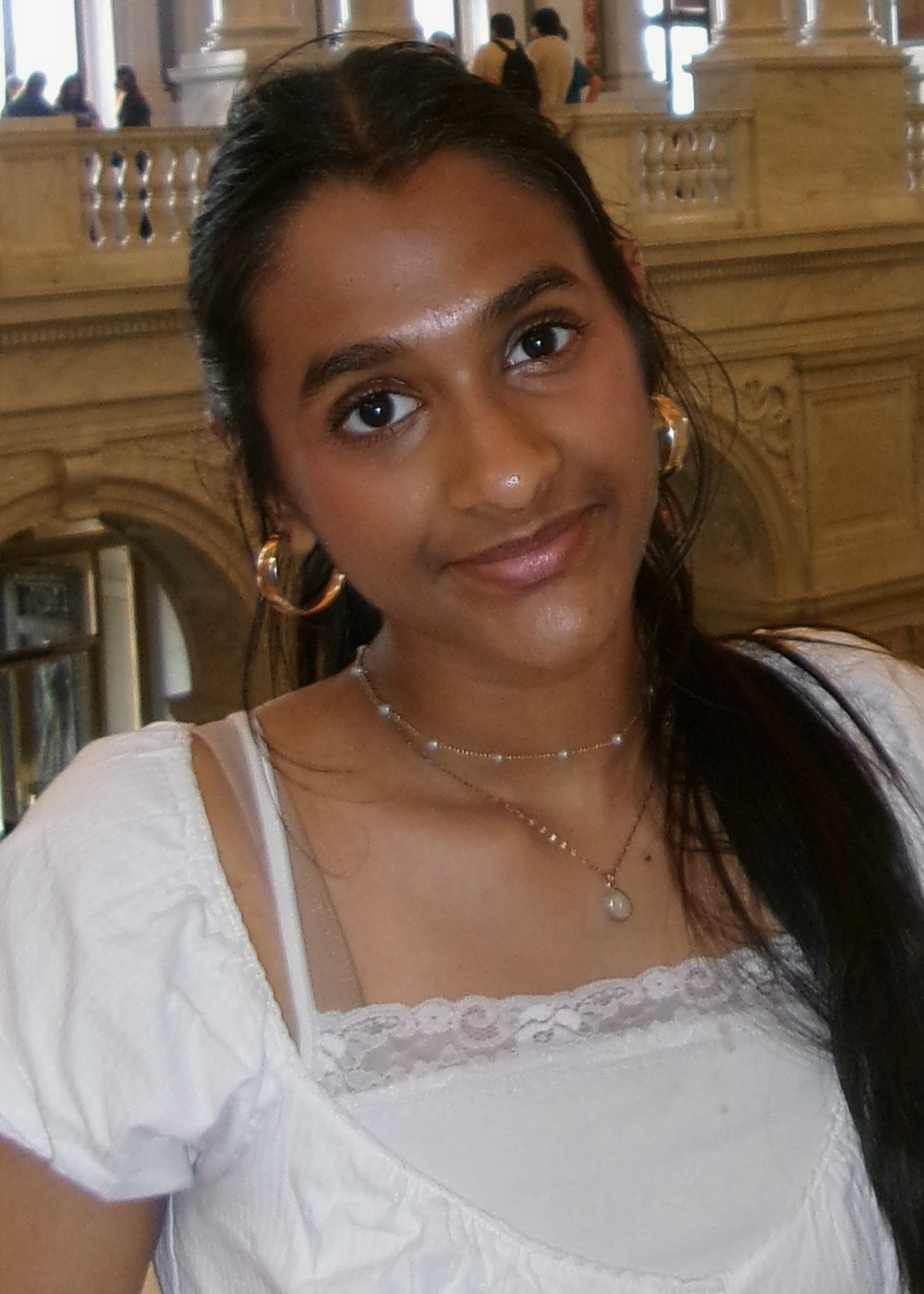 Akshara Krishnan headshot: young woman with long dark hair wearing white shirt poses inside large building